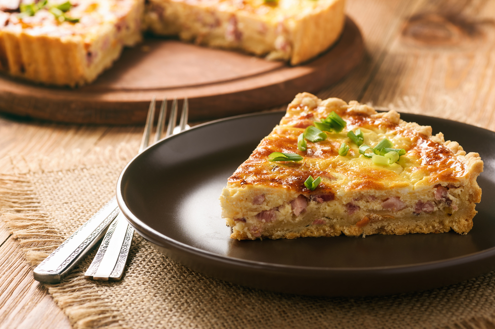 A piece of quiche sits on a dark colored plate, topped with green onions and stuffed with ham.