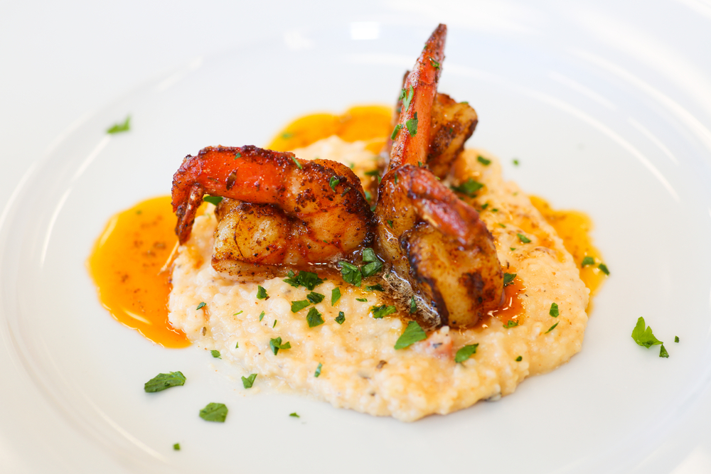 This classic dish of shrimp and grits is displayed on a white plate: the grits are yellow with cheese, and three shrimp sit on tip. The dish is garnish with greens. 
