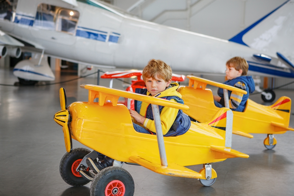 Two young boys in toy airplanes next to a life size plane at Frontiers of Flight, one of the best Dallas kids activities.