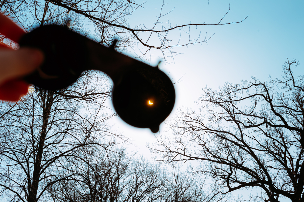 A hand holds up a pair of solar viewers, with a solar eclipse seen in the eyepiece, with trees in the background, similar to how the Arkansas Solar Eclipse will look.