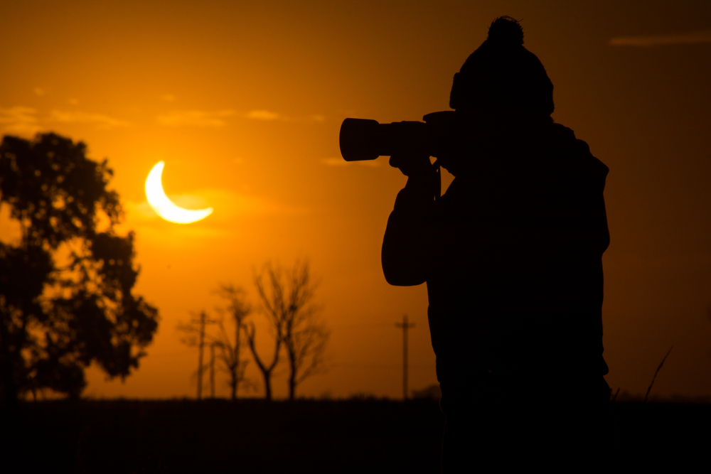 A photographer in a knit hat holds a large camera lens to photograph a solar eclipse against an orange sky.