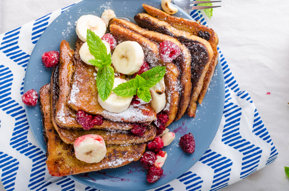 Bananas, berries, and powdered sugar cover 8 slices of French toast on a blue plate with green garnish.