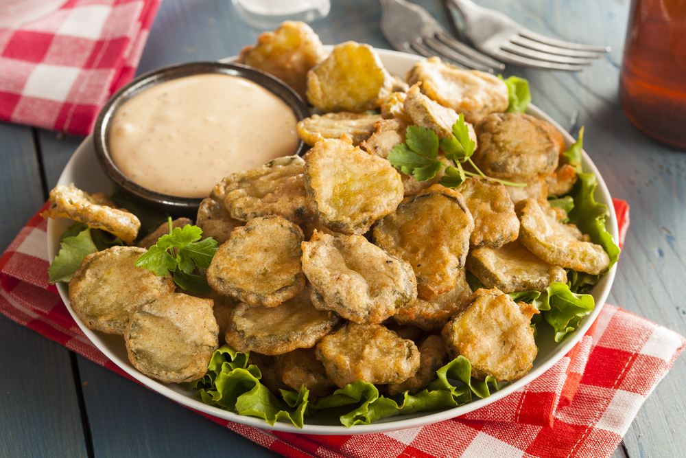 Fried pickle chips sit on a bed of greens with a small dish of dipping sauce, on a red and white checkered napkin
