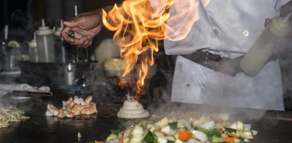 A Japanese chef lights a ring of onions sitting on the grill on fire during a hibachi dinner, like at Ichiban, one of the best restaurants in Roanoke for hibachi.