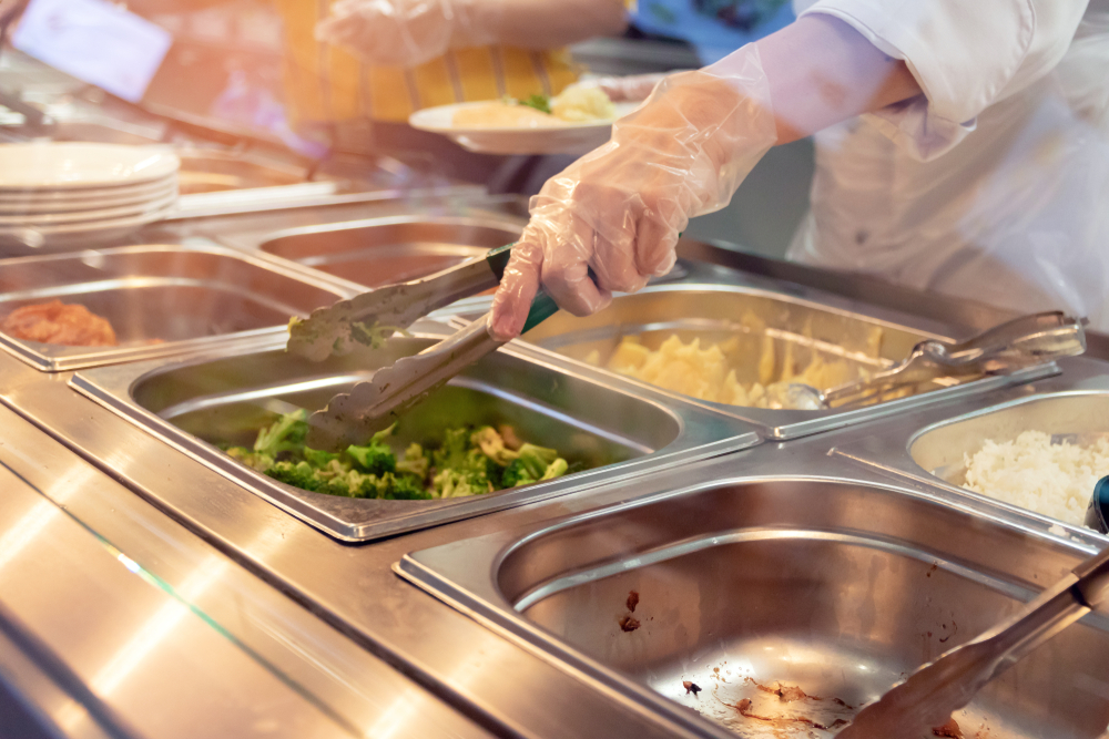 A server's gloved hand holds tongs with which to serve from trays of food at a cafeteria.