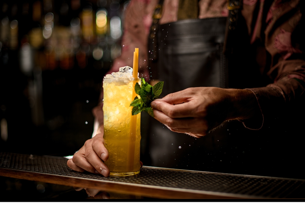 A bartenders hands put a garnish on a tall, amber-colored cocktail.