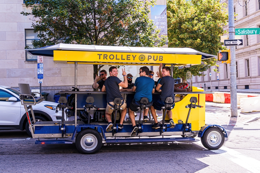 A group of guys on a trolley pub smile and wave as they pedal through downtown and enjoy their drinks. 