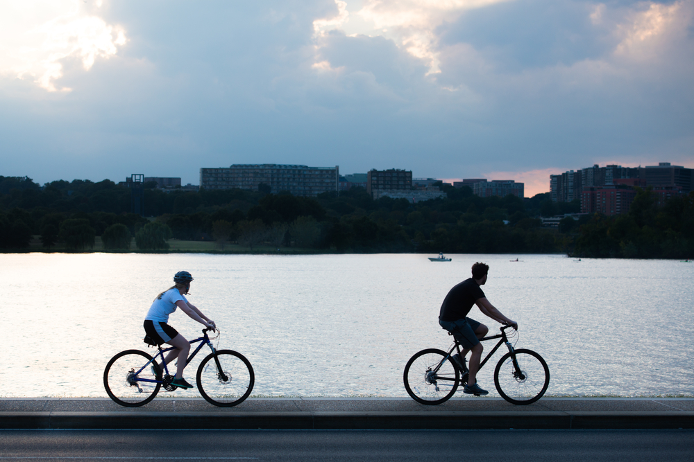 Two bikers circle the cusp of a lake and the edge of a city on a bike tour during sunset or dusk.