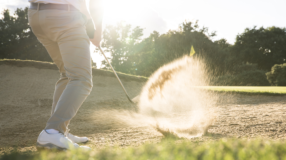 A person's lower half as they swing at a golf ball in a bunker, spraying sand.