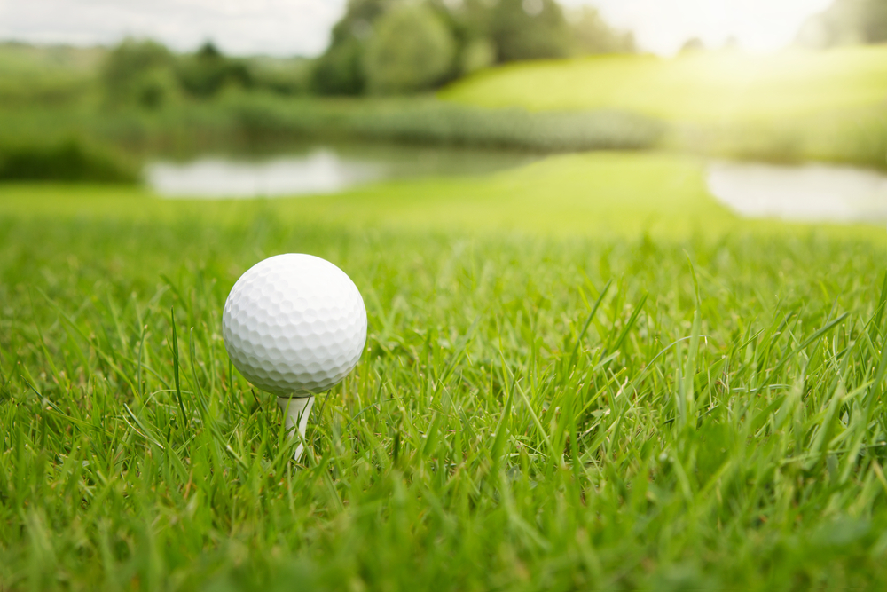 A golf ball sits on a tee, surrounded by green grass, with a strip of land flanked by water in the distance.