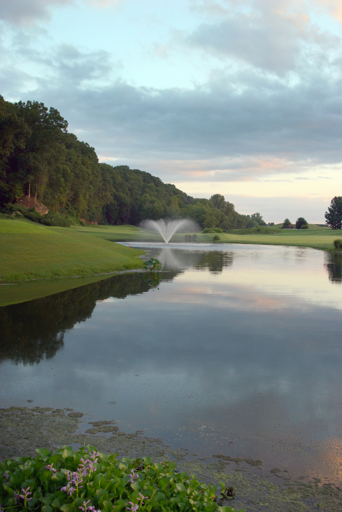 A fountain sprays water in a manmade pond on a golf course at dusk.