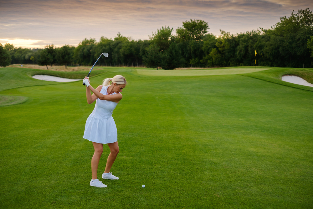A woman in a white outfit on the fairway with a 7 iron driving to the green