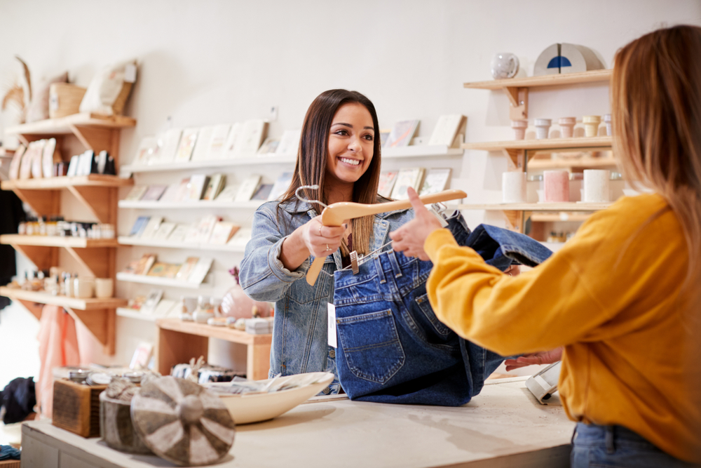 a woman stands behind the counter and a woman stands on the other side of the counter as she purchases a pair of blue jeans