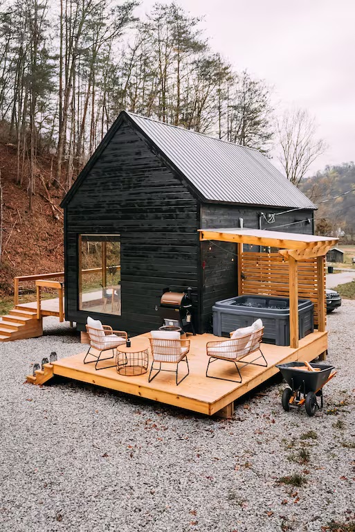 THe stark black wooden cabin with the hot tub under the outdoor pergola with outdoor seating