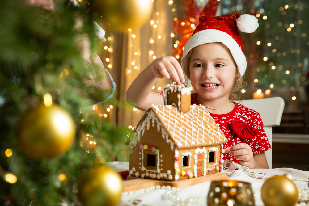a girl decorating a ginger bread house with a big red Santa hat and christmas trees 