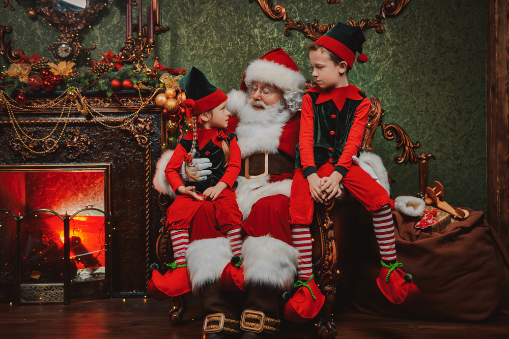 A young boy and girl dressed as elves sitting on Santa's lap in front of a fireplace at North Pole Texas, one of the best places to experience Christmas in Dallas.