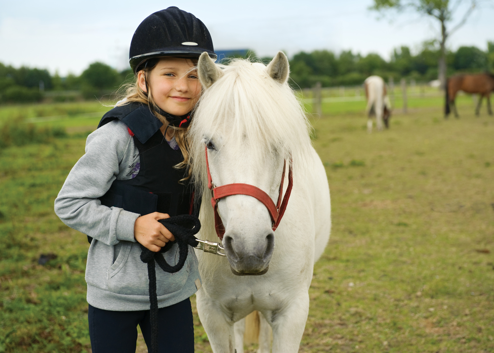 a young child stands next to a pony as she holds its lead rope, shes wearing a helmet and looking at the camera