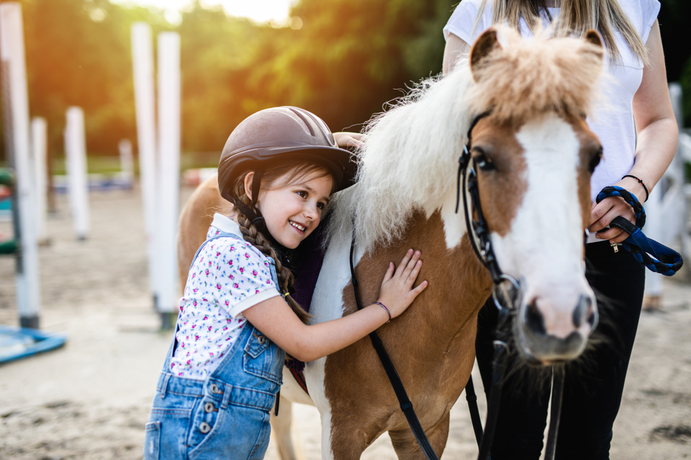 a little girl wearing a helmet standing next to a pony, she is hugging the pony and a woman stands next to them holding the horses lead rope