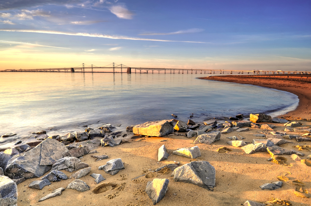 view of the Chesapeake bay bridge from the shore, with large rocks on the beach at sunset