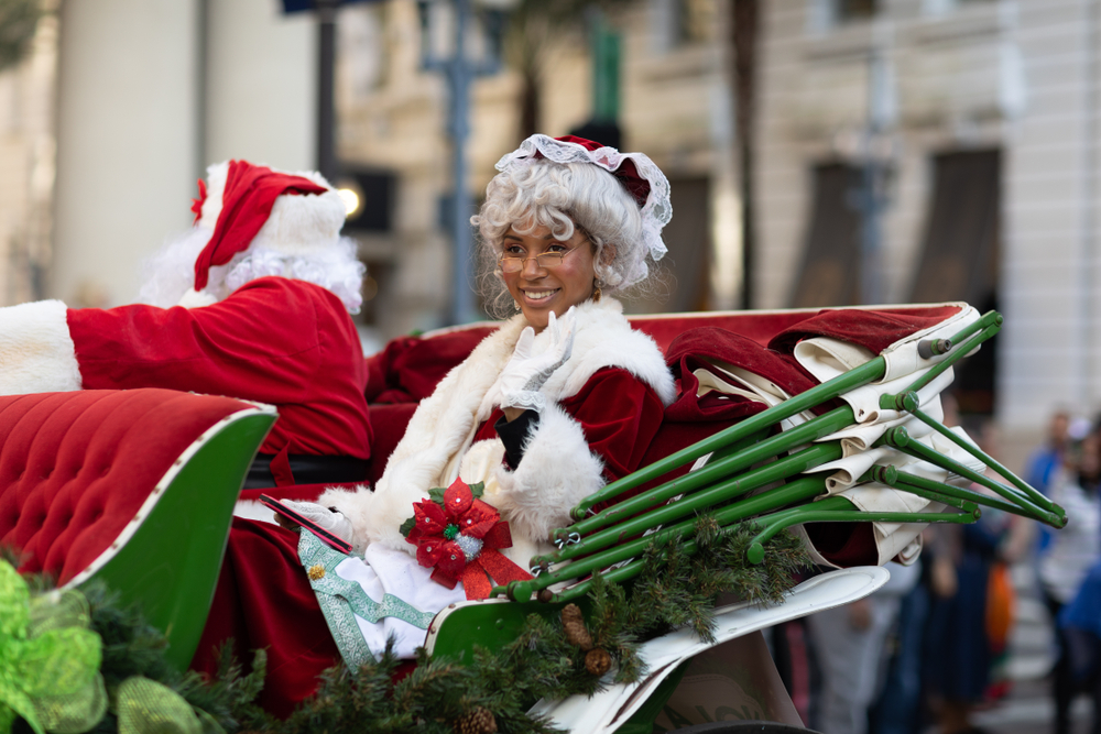santa claus and his wife sit in a sleigh in a christmas parade, mrs claus is looking forward and santa in turned around looking to the side