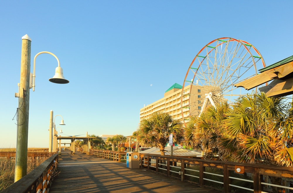 empty boardwalk with a Ferris wheel in the background during the evening, trees lining the boardwalk with lights on the left side 