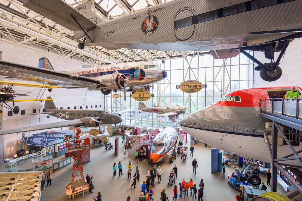 Airplanes displayed in one of the best museums in Washington DC. There are planes hanging from the ceiling and some on the floor and there are people in the museum. 