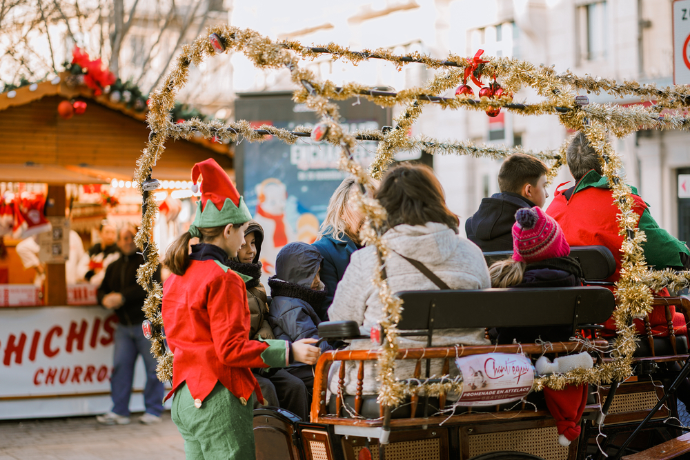 people riding a sleigh decorated with holiday decor like gold tinsel and red ribbons and ornaments, with one person dressed as an elf 