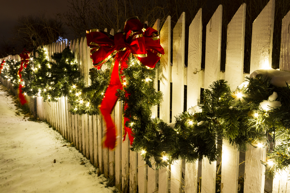 a picket fence in the snow is decorated with garland, christmas lights, and bows