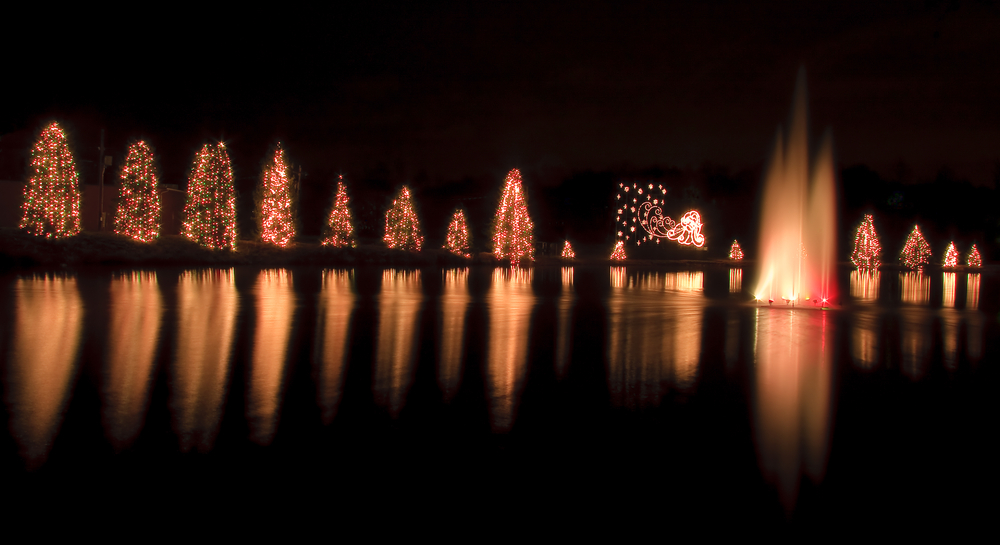 a lake with a fountain in the middle surrounded by trees that have christmas lights on them, mcadenville, christmastown usa