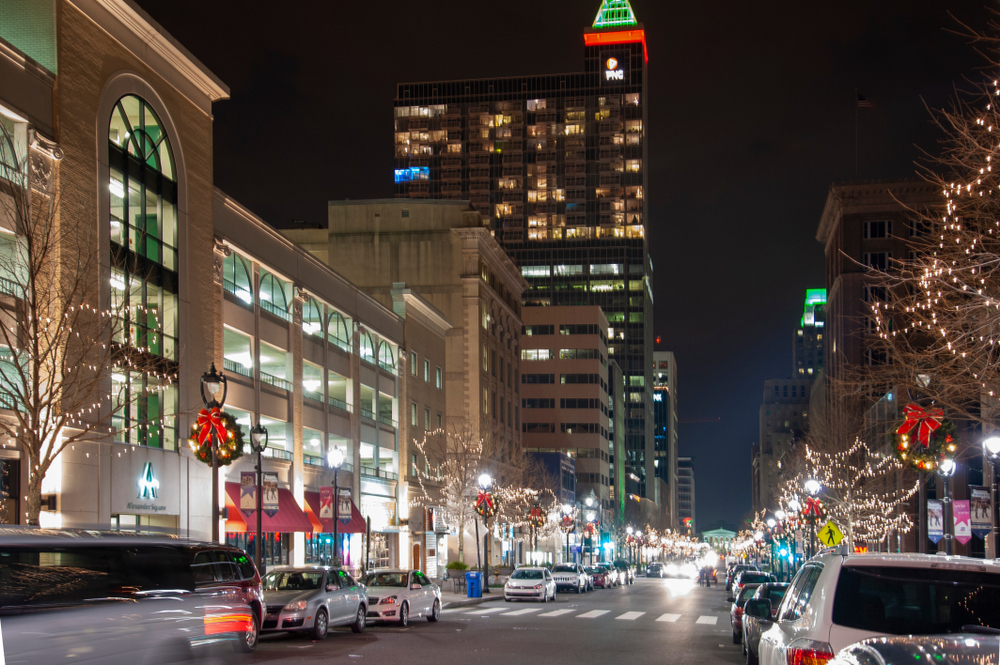 a street decorated with christmas lights and wreaths, there are cars parked along the road and it is night time