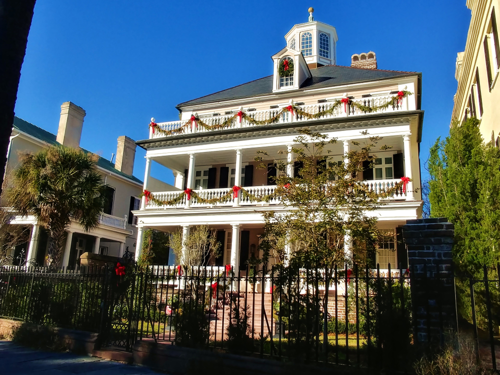 Historical Charleston building with 3 levels of Christmas decorations. The building is decorated in red ribbons and wreaths all across the front of the building, with one singular wreath hanging from the highest window of the building 