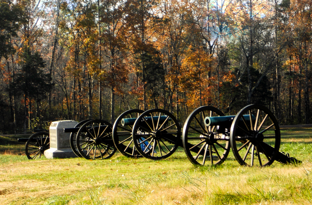 Four Civil war cannons stationed in a field that served as the battlefield for Chickamauga Battle and turned into the National Military State Park in Georgia. The Cannons are surrounded by autumn trees. 