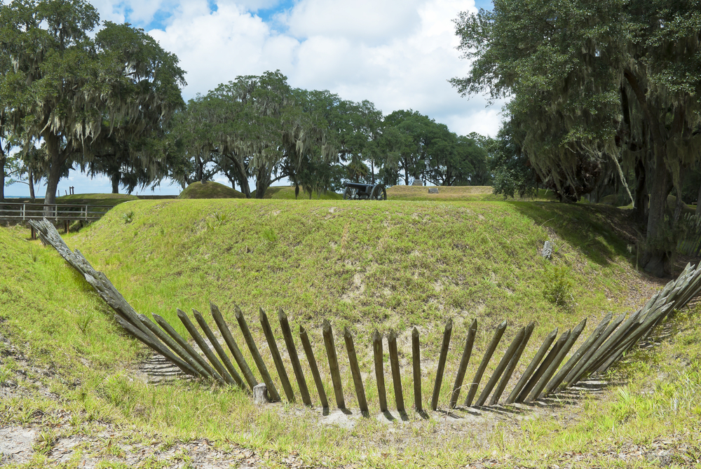 Wooded stake earthwork that makes up the Fort McAllister fortification with a cannon in the background on top of the hill on a sunny day 