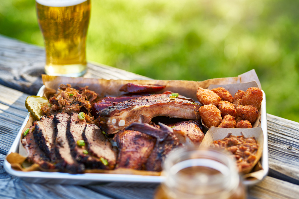 A tray of Texas BBQ, which is one of the best things about living in Texas, sits on a picnic table with a glass of beer next to it, on a sunny day.