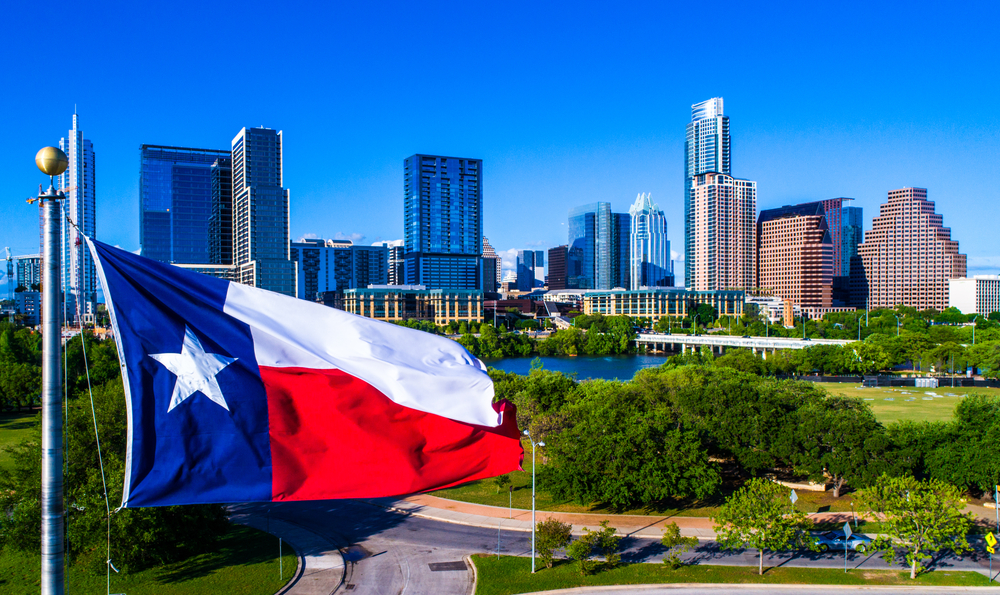 The red, white, and blue flag of Texas waves over the Austin skyline on a sunny day in Texas.