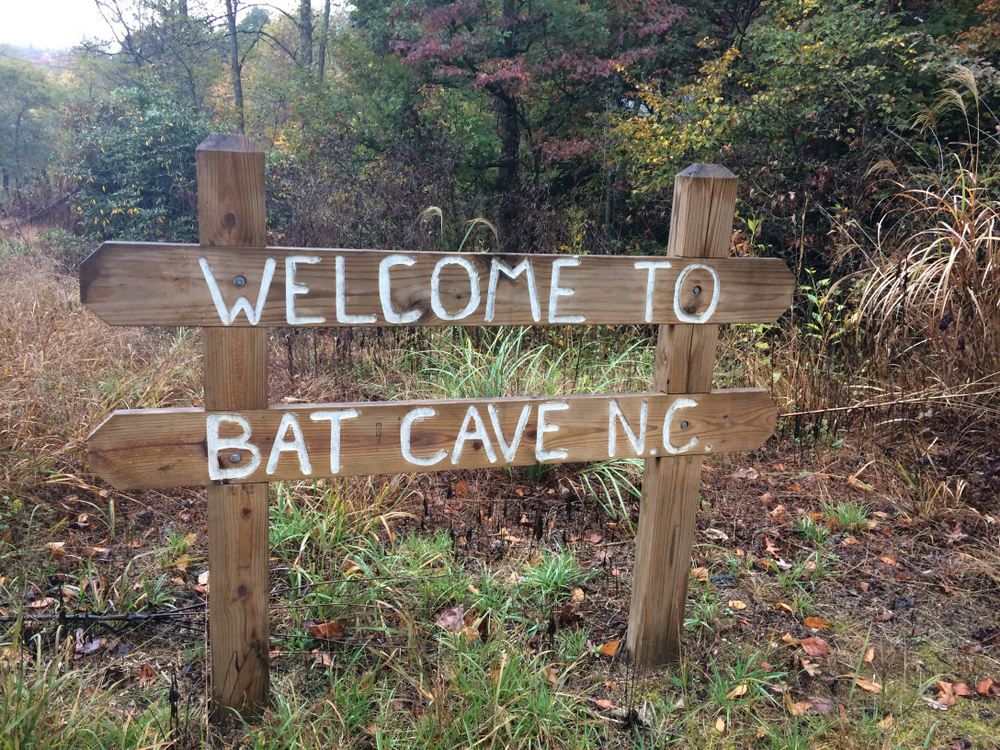 wooden sign with white letters welcoming visitors to bat cave in North Carolina, surrounded by grass and trees with multi-colored leaves 