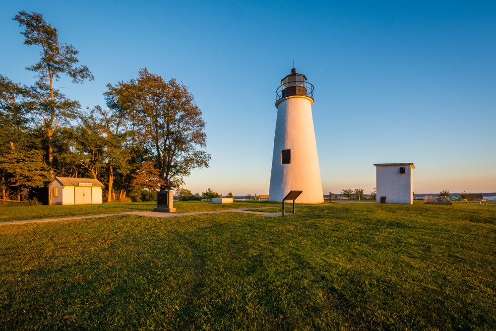 historic turkey point lighthouse at sunrise with a small building to the left and trees in the background, surrounded by green grass  