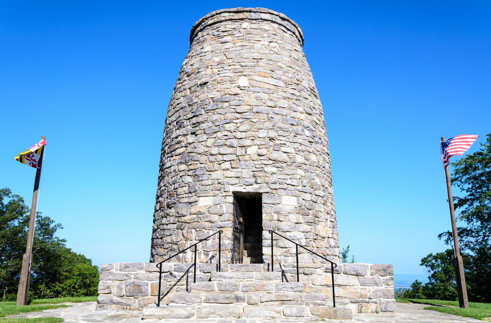 historic stone building located at the Washington monument state park, centered between two flags with the American flag on the right of the photo 