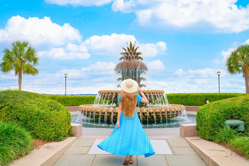 Young woman in flowing blue dress walking toward the Pineapple Fountain in Waterfront Park while holding her sun hat on her head.