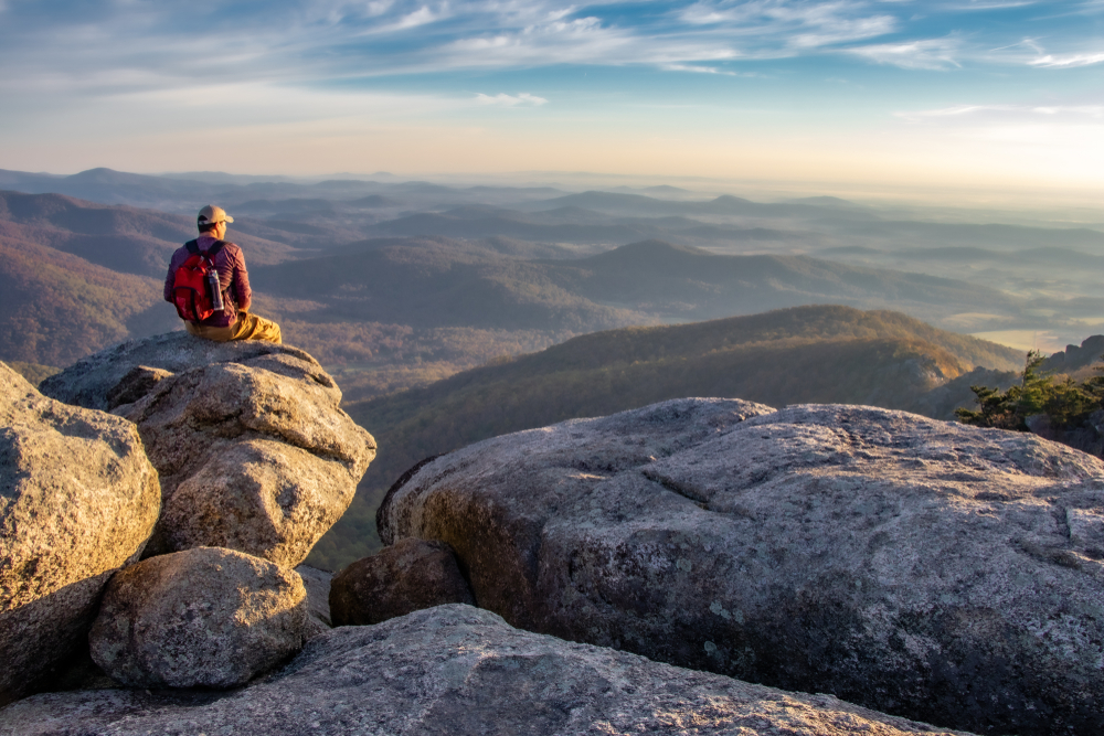 a person sitting on the of the Old Rag Mountain trail after they have completed the trail taking a break. they are looking out at the mountain during sunset 