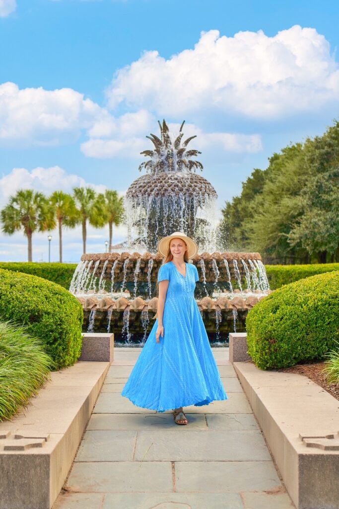 Woman in flowing blue dress and sun hat smiling in front of the Pineapple Fountain in Waterfront Park with manicured hedges.