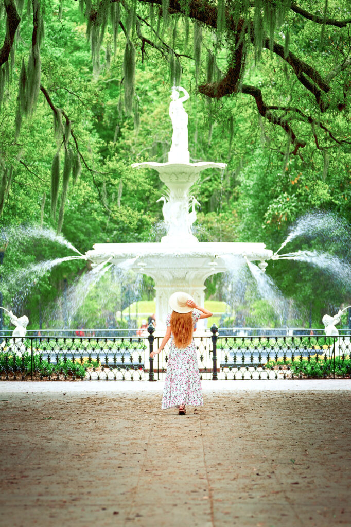 Woman in a sun hat and long, floral white dress walking toward the fountain in Forsyth Park.
