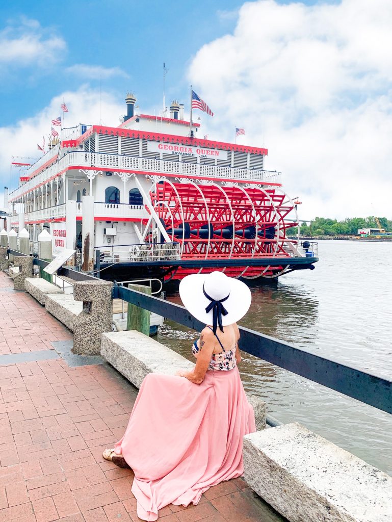 Woman in a pink dress and floppy sun hat sitting next to the river looking at the Georgia Queen River Boat.