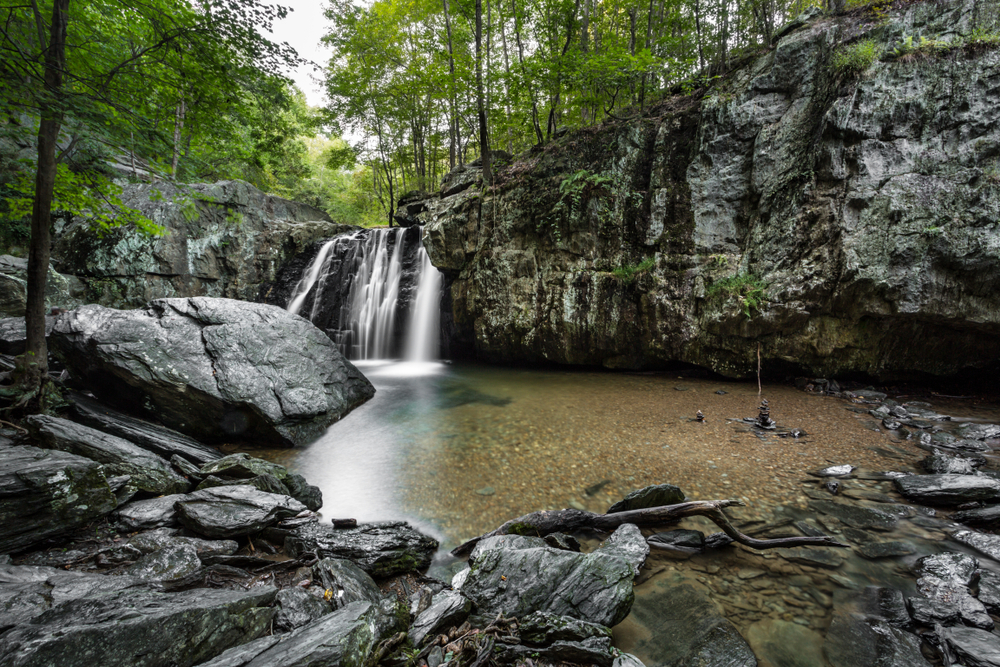 A view of a waterfall over a rocky cliff edge that falls into a crystal clear pool surrounded by lush greenery best waterfalls in Maryland