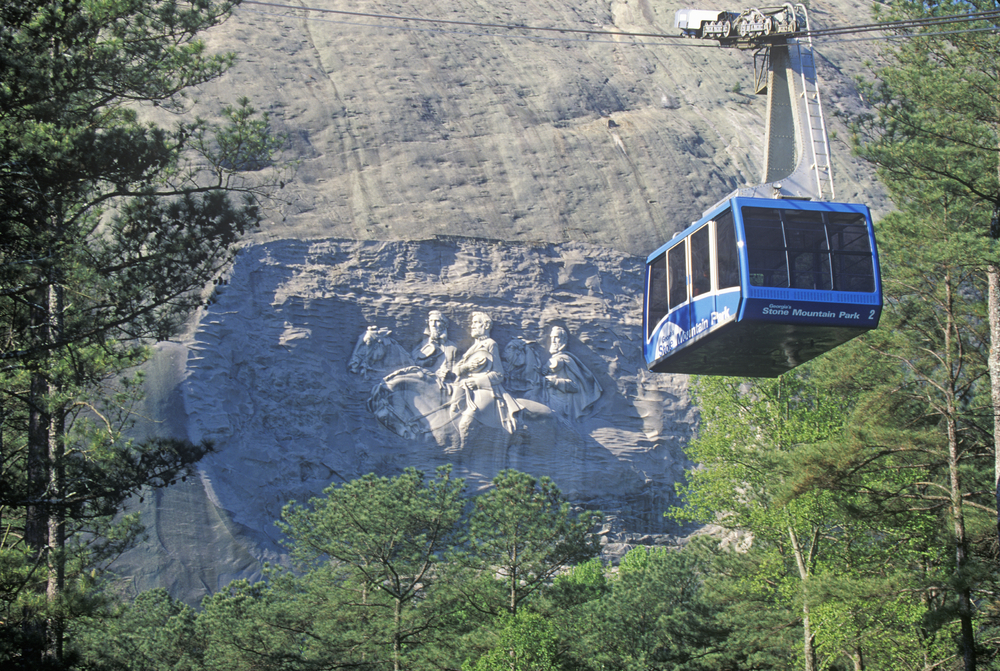 Blue aerial cable car going over trees with a mountain in the background with a carving of men on horses.