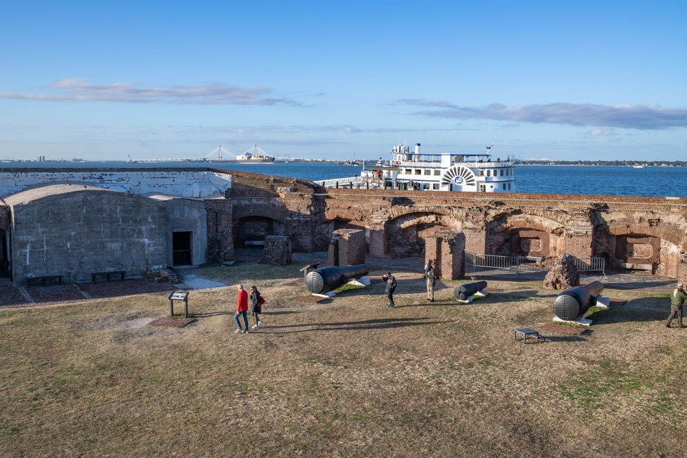 people walking about and looking at the fort during the winter. there is a white ferry in the back round as well as a white bridge and a few smaller boats 