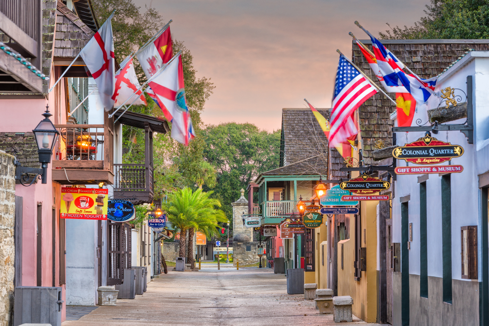 a street with buildings on both sides that are displaying flags of different countries out front, the road is cobbletsones and there are palm trees in the background