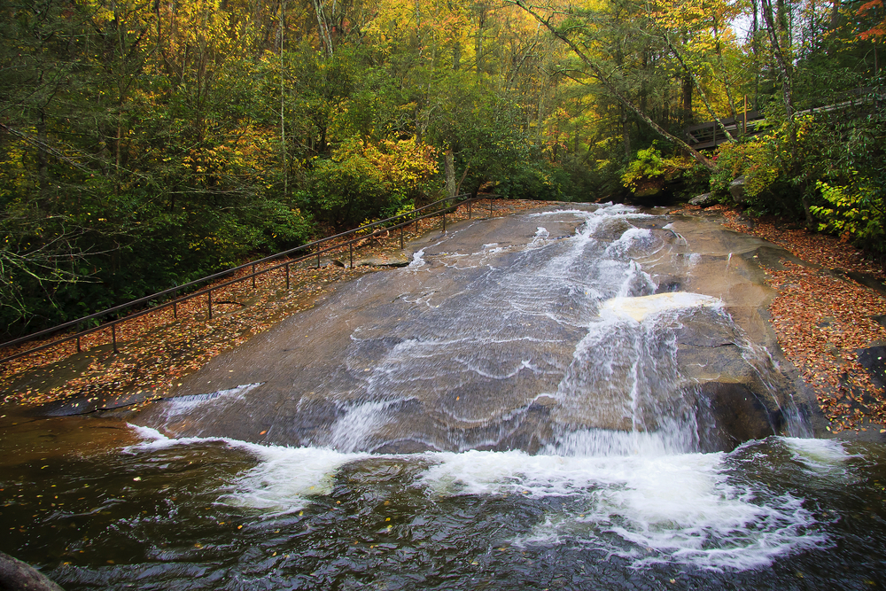 a hand rail on the left going up a sloping natural rock face, a natural waterslide in the middle of the photo, the sliding rock is surrounded by fall foliage and trees