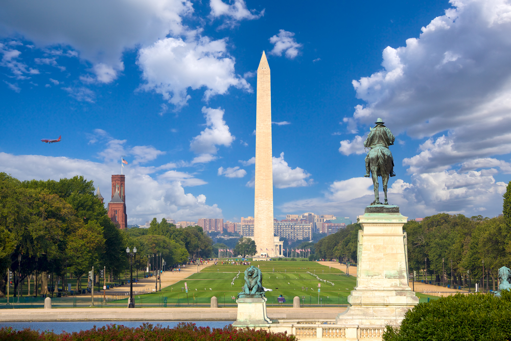 the famous Washington monument stands tall on a bright day, a plan is flying to the left of it and there is a large green grassy area in front of it 