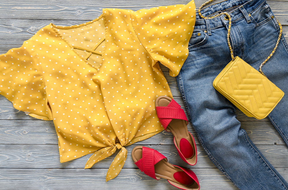 Flat lay image of a yellow, polka dot blouse, jeans, red sandals, and a yellow purse.